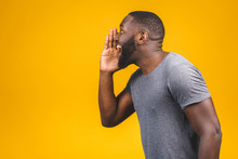 Side View Portrait Of A Young African American Man Screaming Out Loud With Hand At His Mouth Isolated On The Yellow Background.