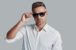 © G-Stock Faces - In his own style. Handsome young man looking at camera and adjusting sunglasses while standing against grey background