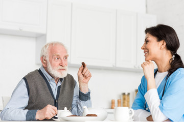 Cheerful grey haired man sitting on kitchen with cookie in hand, and looking at nurse