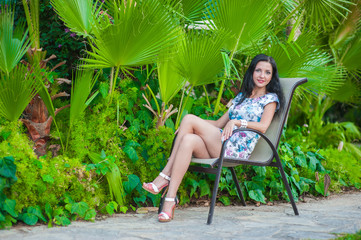 The girl sits near the palm leaves. A beautiful brunette in a dress sits on a chair near the vegetation, resting in the warm lands.