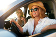 © artiemedvedev - Two young cheerful smiling women in a car on vacation trip to the sea beach. Girl in glasses driving a vehicle from rental on holidays. Girlfriends enjoying summer arrived to ocean shore on holidays.