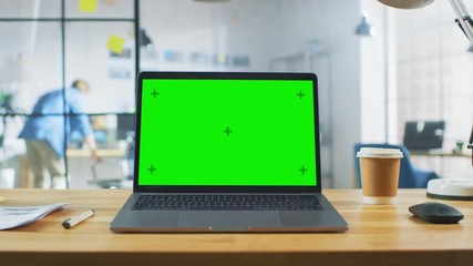 Poster - Laptop on the Desk in the Office Shows Green Mock-up Screen. In the Background Bright Creative Open Space Loft Office HUB with Professional Working
