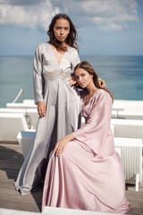 two Beautiful young women models in a long silk pink and gray dresses against the blue sky and sea view. fashion posing. one girl is sitting, other is standing. girls are hugging