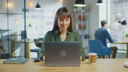 Poster - Young Beautiful Brunette Works on a Laptop Computer in Cool Creative Agency in a Loft Office. She has a Take-away Coffee and a Notebook on Her Table. She Smiles and Laughs. 