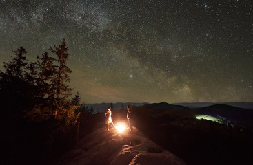 Happy couple hikers having a rest together, standing beside campfire on the top of big boulder in the mountains at night. On background beautiful night starry sky full of stars and Milky way.