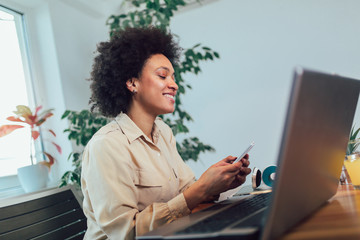 Smiling young African female entrepreneur sitting at a desk in her home office working online with a laptop