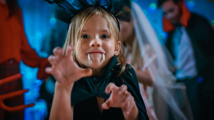 Halloween Costume Party: Cute Little Bat Girl with Sharp Fangs Makes Scary Funny Faces. In the Background Monster Party in Decorated Room and Disco Lights