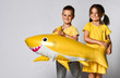 © FAB.1 - Positive young children hold a balloon in the shape of a yellow shark fish, celebrate the holiday, smiling widely, stand on a light background, are in a good mood