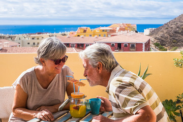 Retired people lifestyle with cheerful senior couple having fun together at home on the terrace drinking from the same glass a juice fruit - city and ocean view for vacation concept