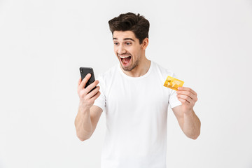 Excited happy young man posing isolated over white wall background using mobile phone holding credit card.