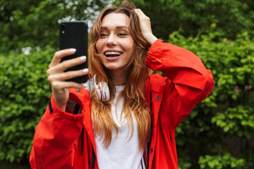 Canvas Print - Image of pretty young woman holding cellphone while walking through green park under rain