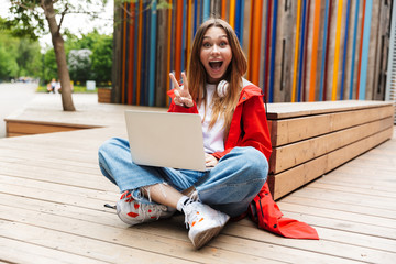 Poster - Beautiful young happy pretty woman in raincoat posing outdoors using laptop computer showing peace gesture.