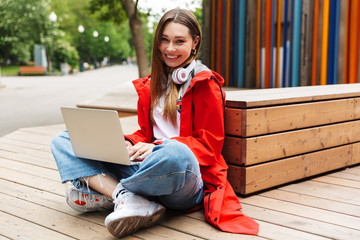 Poster - Beautiful young happy pretty woman in raincoat posing outdoors using laptop computer.