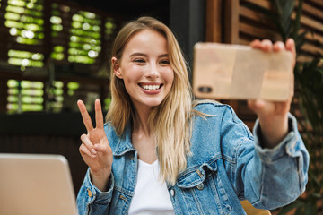 Wall Mural - Happy young pretty blonde woman in cafe using laptop computer and mobile phone take a selfie.