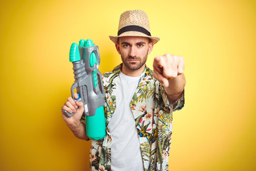 Canvas Print - Young man wearing hawaiian flowers shirt holding water gun over yellow isolated background pointing with finger to the camera and to you, hand sign, positive and confident gesture from the front