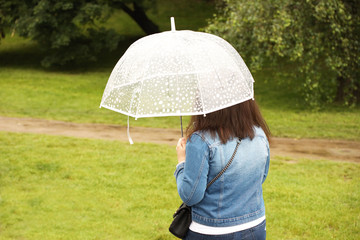 A girl in a denim blue jacket with a white transparent umbrella walks in the rain among the green. Romantic mood. Melancholic wet weather. Walk in rainy weather with a trendy gadget. Autumn-spring