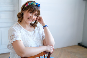 A young plus size woman sits on a chair in a spacious white room. Smile on his face, flirtatious look. Happiness and beauty in any weight. A girl in a long skirt, sandals with high heels