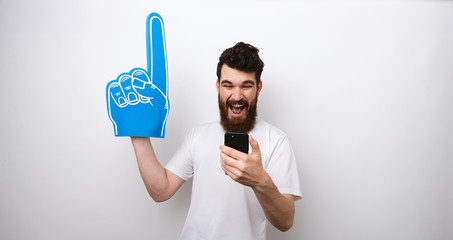 Young bearded man holding a blue foam finger and watching his phone.