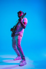 Wall Mural - Young african-american musician playing the guitar like a rockstar on blue studio background in neon light. Concept of music, hobby. Joyful attractive guy improvising. Retro colorful portrait.