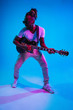 © master1305 - Young african-american musician playing the guitar like a rockstar on blue studio background in neon light. Concept of music, hobby. Joyful attractive guy improvising. Retro colorful portrait.