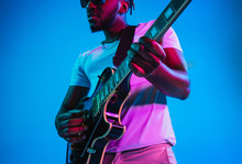 Young African-american Musician Playing The Guitar Like A Rockstar On Blue Studio Background In Neon Light. Concept Of Music, Hobby. Joyful Attractive Guy Improvising. Retro Colorful Portrait.