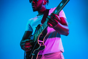 Young african-american musician playing the guitar like a rockstar on blue studio background in neon light. Concept of music, hobby. Joyful attractive guy improvising. Retro colorful portrait.