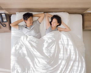 Top view of happy Asian couple smiling, and sleeping together on bed in love and sex concept in a modern bedroom with white blanket in the morning.