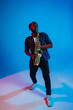© master1305 - Young african-american jazz musician playing the saxophone on blue studio background in trendy neon light. Concept of music, hobby. Joyful attractive guy improvising. Retro colorful portrait of artist
