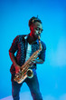© master1305 - Young african-american jazz musician playing the saxophone on blue studio background in trendy neon light. Concept of music, hobby. Joyful attractive guy improvising. Retro colorful portrait of artist