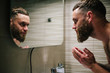 © San4ezz007 - Young bearded hipster man washing his face with water in the bathroom while looking himself in the mirror.