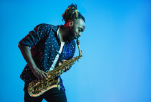 Young African-american Jazz Musician Playing The Saxophone On Blue Studio Background In Trendy Neon Light. Concept Of Music, Hobby. Joyful Attractive Guy Improvising. Retro Colorful Portrait Of Artist
