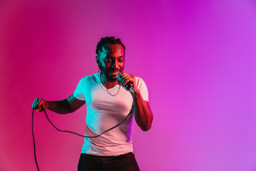 Wall Mural - Young african-american jazz musician with microphone singing a song on purple studio background in trendy neon light. Concept of music, hobby, inspirness. Colorful portrait of joyful attractive artist