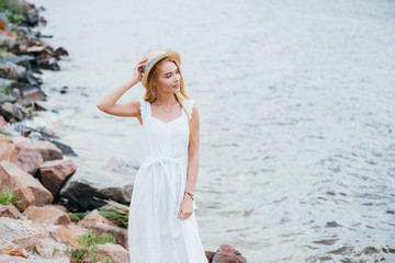 Wall Mural - happy young blonde woman touching straw hat and looking at sea