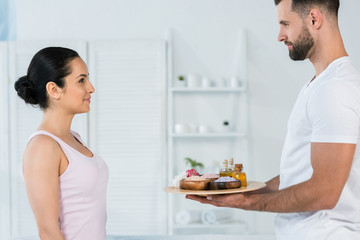 Wall Mural - handsome masseur holding tray with sea salt and oil in bottles near attractive woman