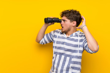 Wall Mural - Blonde man over yellow wall and looking in the distance with binoculars