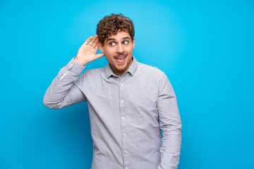 Wall Mural - Blonde man over blue wall listening to something by putting hand on the ear