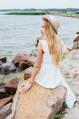 Wall Mural - beautiful blonde woman in straw hat sitting on stones near sea