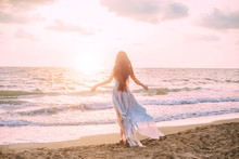 Mysterious Mermaid With Long Black Hair Slowly Walks In Water Of Ocean, Sea Nymph Listens To Wind Like Wave In Long Blue Dress With Flying Train, Looks At Divine Sunset. Photo From The Back, No Face