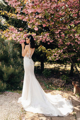 beautiful woman with dark hair in luxurious wedding dresses with accessories posing in garden with blossoming sakura trees