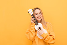 Cute Girl Hugs Ukulele With Her Eyes Closed And Smiles, Isolated On A Yellow Background, Wearing Orange Clothing. Portrait Of A Happy Girl With Hawaiian Guitar In Her Hands. Copy Space