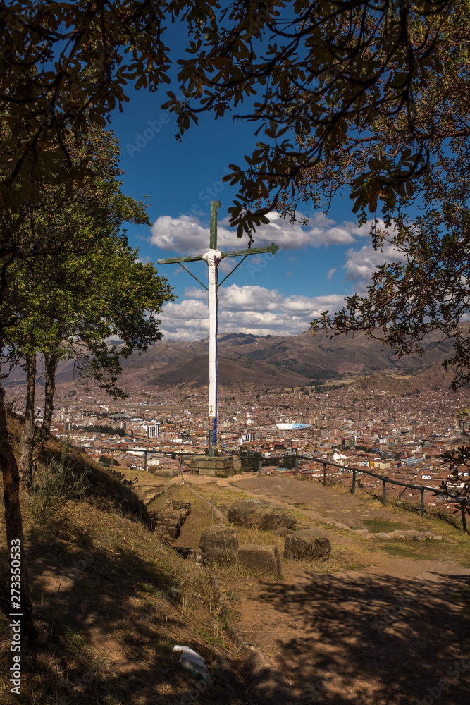A sweeping view of the city of Cusco, the ancient capital city of the Inca empire, the orange clay roofs stand out against the mountains, a cross in the foreground