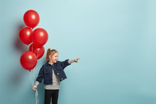 Mirthful Red Haired Little Girl With Pony Tail, Holds Big Bunch Of Red Balloons, Points Directly Into Distance, Wears Striped Jumper And Denim Jacket Shows Direction Somewhere Being On Childrens Party