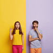 © wayhome.studio - Shocked woman points upwards, keeps mouth opened, thoughtful unshaven man holds chin, stand shoulder to shoulder, pose over purple and yellow background, free space for your promotional content
