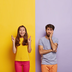 Happy woman in casual clothes, keeps fingers crossed, believes in good luck, sad dejected man feels bored, stands near, pose against yellow and purple striped background. Body language concept