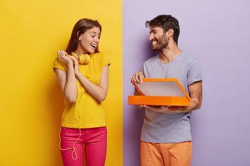 Wall Mural - Happy young male model opens box, makes surprise for girlfriend on birthday, shows something in package, isolated over purple background. Glad lovely woman with headphones on neck, receives gift