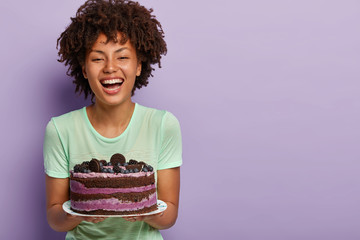 Wall Mural - Happy birthday girl laughs joyfully, holds big tasty fruit cake, likes eating sweet food, improves mood with raising sugar in blood, has positive face look, stands over purple background, blank space