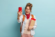 Smiling good looking woman makes selfie via cell phone, being in high spirit, enjoys free time after classes, wears red bandana around neck, rucksack on back, holds spiral diary, isolated on blue wall
