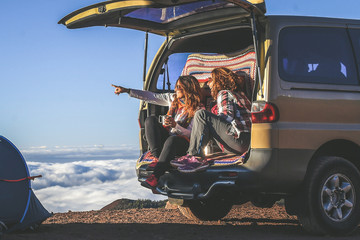 Couple of woman enjoying outdoor sitting on the back of a 4x4 van. Gorgeous mountain panorama on the background. Two happy girls toast with cups of coffee. Tourists enjoy a beautiful vacation together