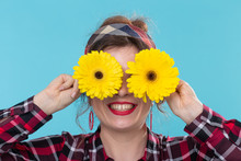 Close-up Positive Smiling Young Woman In A Red Checkered Shirt Holding Yellow Flowers On Her Eyes Posing Against A Blue Background. Concept Incognito And Fun.