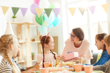 Multi ethnic group of children celebrating birthday sitting at table in cafe, focus on mother giving present to daughter, copy space